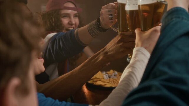 Side view closeup slowmo shot of cheerful young diverse friends wearing team jersey clinking beer glasses sitting at bar counter and having fun together watching sports game