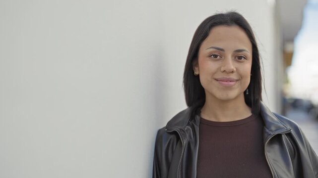 Young hispanic woman smiles and leans against a pale wall wearing a leather jacket and brown sweater on a street; confidence.