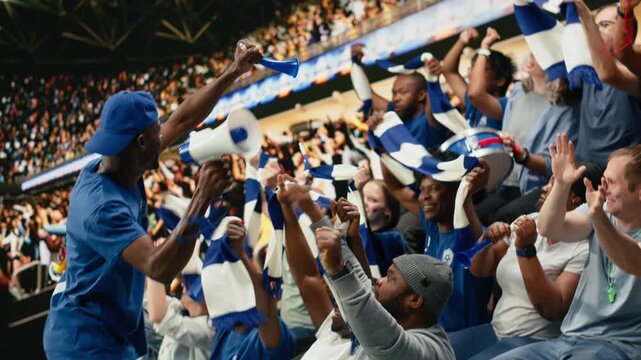Blue team football capo standing above crowd with megaphone, leading passionate supporter section during energetic stadium chant, organized fans producing powerful support chants with crowd energy.