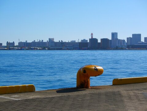 A yellow mooring bollard on the pier at Kobe Mosaic with a wide view of the blue harbor water and city skyline on November 23 2025 in Kobe Hyogo Japan