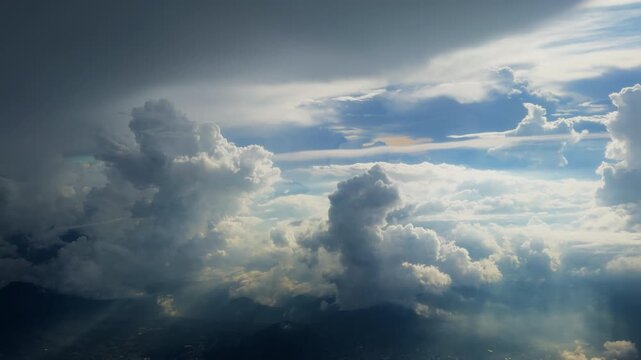 Breathtaking aerial view of massive cumulus clouds towering above dark mountain silhouettes with golden sunlight rays piercing through dramatic storm formations in a moody atmospheric sky.