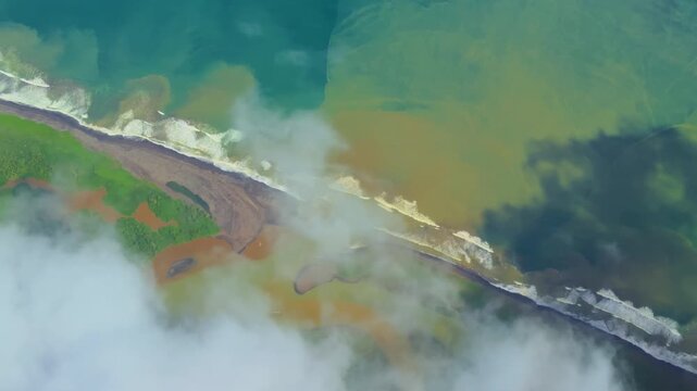 Stunning aerial footage of tropical coastline where turquoise ocean meets lush green vegetation. White clouds drift over sandy beaches and shallow waters with vibrant color gradients.
