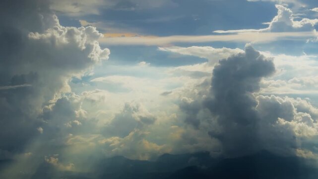 Majestic cumulus clouds tower over distant mountain silhouettes as golden sunlight rays pierce through the dramatic sky. Soft light illuminates cloud layers creating depth and atmosphere.