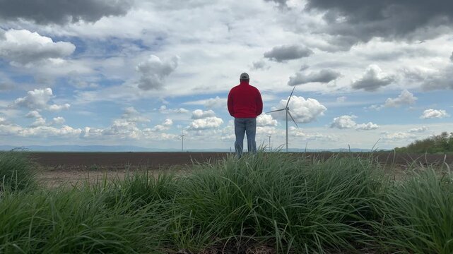 Cinematic footage of an adult man standing on a rural roadside overlooking a wind turbine and open agricultural landscape beneath a dramatic sky filled with towering clouds. 