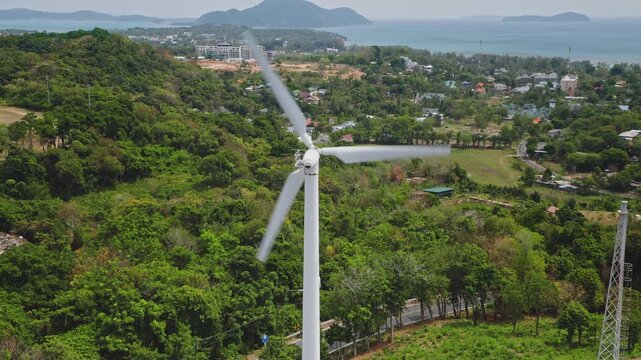 Wind turbine spinning its blades, generating renewable electricity above a lush green tropical landscape with coastal towns and ocean in the background on Phuket, Thailand. Aerial view drone shot