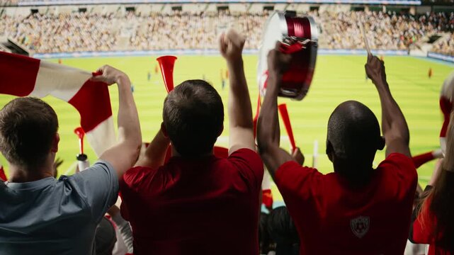 Back view of soccer supporters in red jerseys watching the field from the stands, waving scarves during live football match celebration.