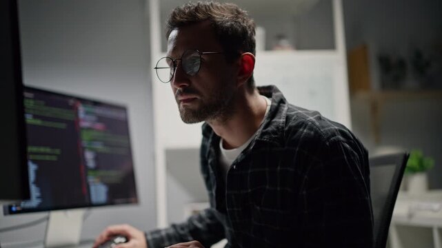 A professional man programmer using a mouse, typing on a computer keyboard, and looking at the computer code screen at night.