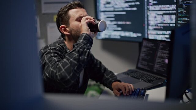 Male web developer drinking coffee while working late on computer, tired application programmer coding software on laptop at night office.