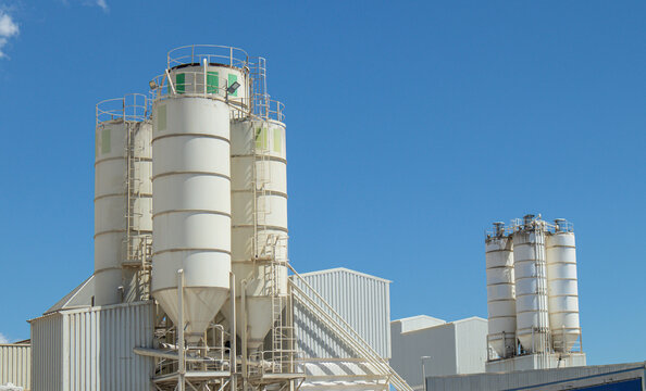 Concrete mixing plant silos and storage tanks stand tall against a clear blue sky in an industrial area with no visible activity