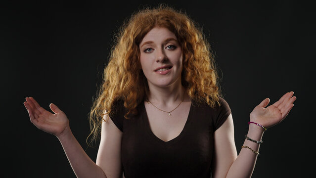 Young woman with red hair raising hands, unsure and hesitant on dark background