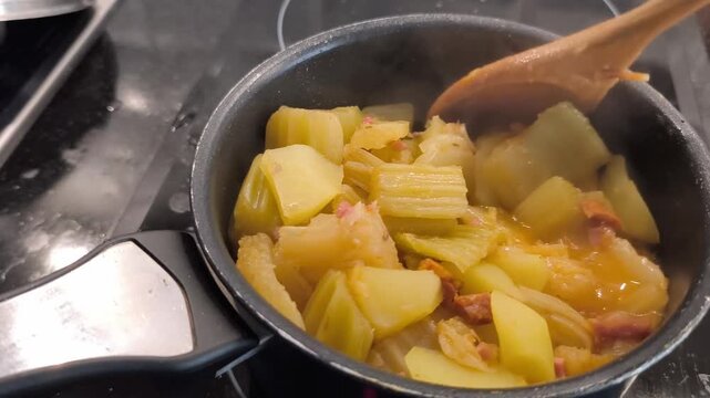 Close up of a person stirring a steaming cardoon and potato stew with a wooden spoon in a pot