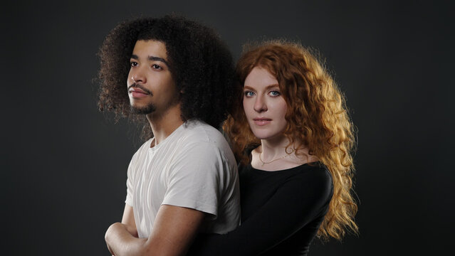 Diverse couple standing close together, woman embracing man from behind, looking at camera