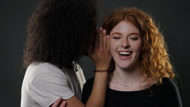 Diverse couple sharing a secret, man whispering in woman's ear, both smiling and laughing