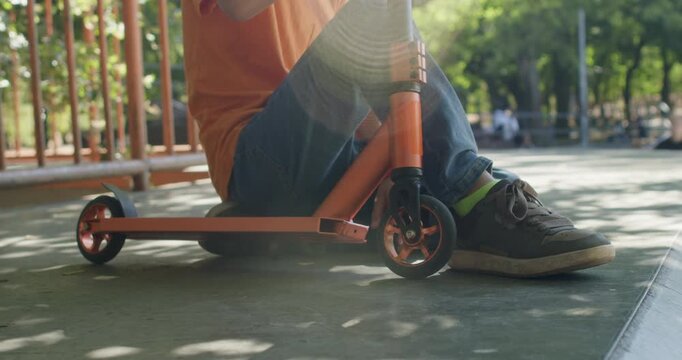 Boy rides orange scooter back and forth while sitting low on deck. Child pushes with shoes rocking front wheel along smooth concrete surface near railing