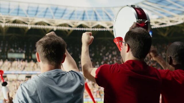 Back view of passionate soccer supporters in red jerseys cheering in packed stadium during live match with supporter chants and stadium atmosphere.