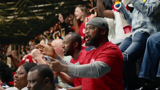 Shocked black fan celebrating betting lucky win with adrenaline, jumping with his friends after final whistle inside the stadium.