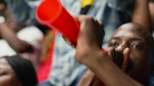 Close side view of passionate male football fan blowing red vuvuzela in stadium stands during intense live soccer match, diverse multigenerational supporters in the fan section, team spirit.