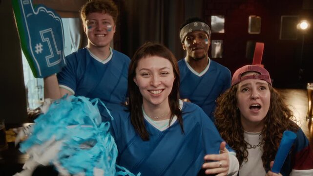 POV shot of excited young diverse sports fans in team apparel looking at camera while cheering with foam hands and cheerleading pom pom watching live game in bar