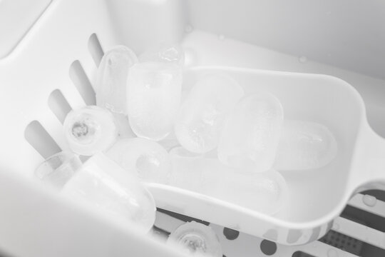 Overhead view inside modern white ice maker showing basket filled with clear bullet ice cubes and plastic scoop. 