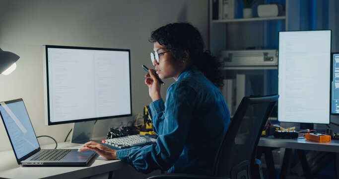 Young African American female programmer coding AI software on multiple screens in night office. Network security, cybersecurity, machine learning, working late, overtime, robotics automation concept.