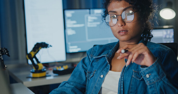 Close-up young Black female programmer coding AI software on multiple screens in night office. Network security, cybersecurity, machine learning, working late, overtime, robotics automation concept.