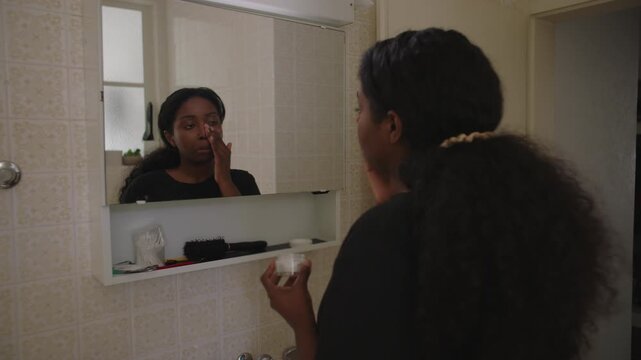 African American woman applying hydrating cream to face while looking at her reflection in bathroom mirror creating calm and attentive self care moment in home interior