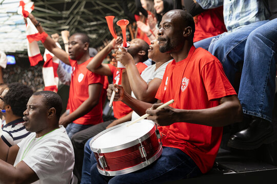 Passionate football supporter playing red fan drum in stadium stands while crowd waves scarves and blows vuvuzelas. Diverse football ultras create intense crowd noise filled with excitement.