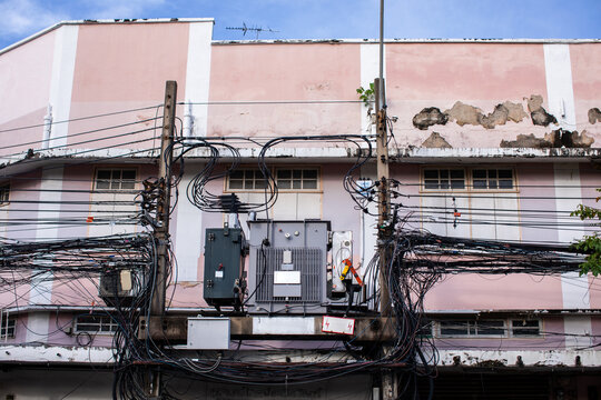 A large electric transformer mounted on concrete poles, tangled in a messy web of overhead power and telecom cables against a weathered pink building facade.