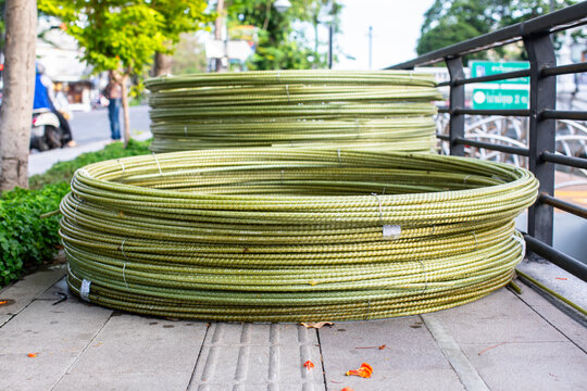 Coiled coils of greenish fiberglass rebar (GFRP) stacked on a city sidewalk. The lightweight, ribbed composite material is bundled with wire, showing a modern alternative.