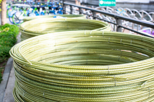 Coiled coils of greenish fiberglass rebar (GFRP) stacked on a city sidewalk. The lightweight, ribbed composite material is bundled with wire, showing a modern alternative.