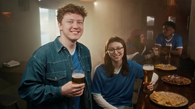 Medium portrait of young Caucasian man and young biracial girl wearing blue team jersey sitting at bar counter with assorted snacks and clinking beer glasses while smiling at camera
