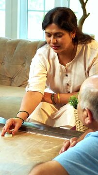 Playing carom or carrom tabletop game together, Indian family of four with senior parents and young adult son and daughter enjoying fun bonding time in cozy living room at home indoor leisure activity