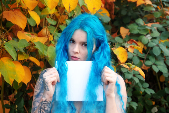 Portrait of a woman with vivid blue hair standing before colorful autumn foliage while holding a blank card creating a striking and expressive outdoor scene
