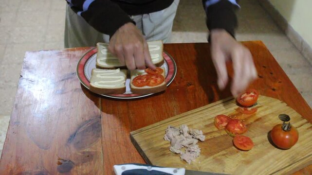 Hombre coloca rodajas de tomates frescos sobre rebanadas de pan lactal untadas con mayonesa durante la preparaci&oacute;n de s&aacute;ndwiches caseros en la cocina. La escena muestra un momento cotidiano de cocina.