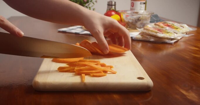 Close up of hands slicing fresh carrot into thin strips with a kitchen knife on a wooden cutting board during vegetable preparation for home cooking.