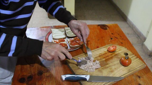 Hombre desmenuzando at&uacute;n con dos tenedores para colocarlo sobre rodajas de tomate en un s&aacute;ndwich fresco preparado sobre una tabla de madera de cocina. La escena muestra ingredientes naturales.