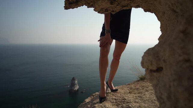 Legs, cliff, sea. Slender female legs in black high heels and a short dress standing dramatically on a rocky cliff edge overlooking the blue ocean.
