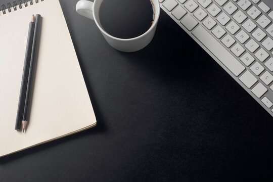 Top view of a white mug filled with black coffee placed on a dark workspace next to a computer keyboard, pencils, and an open spiral notebook. Minimalist conceptual backdrop for remote work, business 