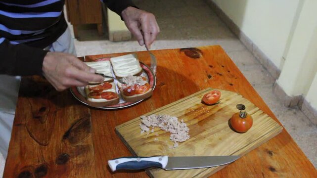 Hombre colocando picadillo de at&uacute;n sobre rodajas de tomate en pan lactal de miga para preparar un s&aacute;ndwich casero en la cocina. Escena culinaria dom&eacute;stica con ingredientes frescos.