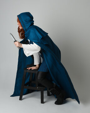 full length portrait of female model wearing medieval fantasy warrior historical costume with blue hooded cloak. crouching seated pose, holding knife dagger weapon, silhouette figure on studio backgro