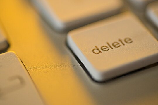 Extreme macro close-up of the "delete" key on a computer keyboard illuminated by warm golden light. Conceptual backdrop for data removal, digital erasing, privacy protection, system troubleshooting