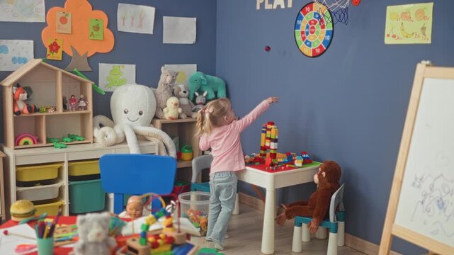 Little blonde girl stacks colorful blocks at a plastic play table amid plush toys and shelving in a preschool building; curiosity wonder imagination play.