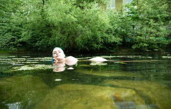 Blue haired woman floating in clear green river surrounded by reflections and water plants creating surreal natural portrait with tranquil mysterious atmosphere