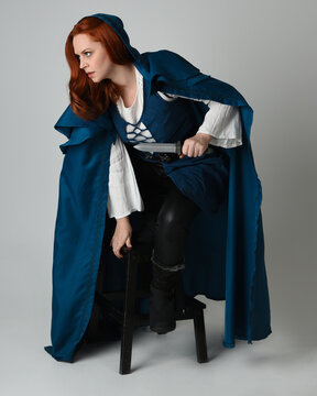 full length portrait of female model wearing medieval fantasy warrior historical costume with blue hooded cloak. crouching seated pose, holding knife dagger weapon, silhouette figure on studio backgro
