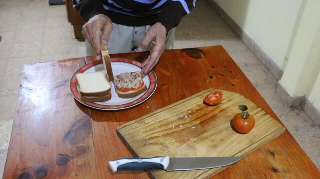 Hombre terminando de preparar s&aacute;ndwiches de pan lactal de miga en la cocina, colocando las tapas de pan sobre tomates frescos, at&uacute;n y mayonesa. Escena culinaria casera con ingredientes frescos.