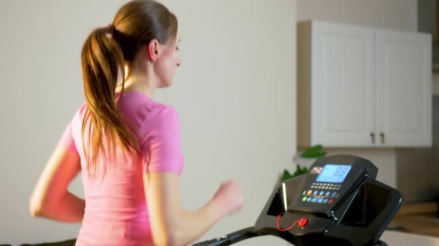 Rear view woman running on treadmill during home cardio workout. Female athlete training on fitness machine while following running program on digital display. Indoor endurance exercise routine