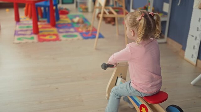 Little blonde girl rides wooden tricycle toward a red play table in a preschool classroom building; playful childhood.