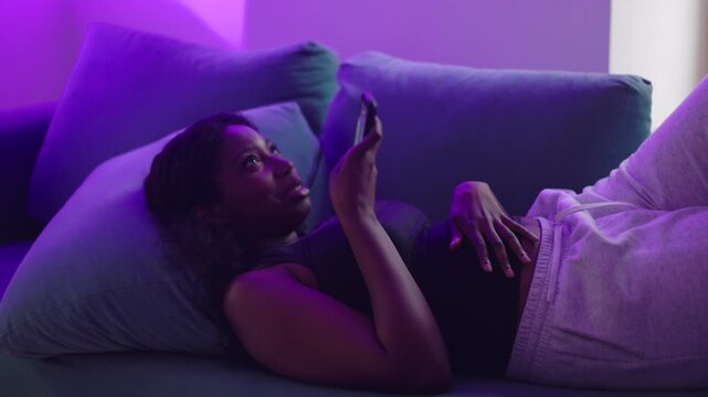 African American woman reclining on couch recording voice message on smartphone, speaking casually while relaxed, capturing everyday communication and modern digital lifestyle at home