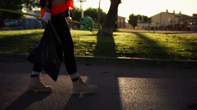 Unrecognizable municipal worker in reflective vest walking through public park carrying rake and garbage bag, supporting seasonal city maintenance and outdoor cleaning.