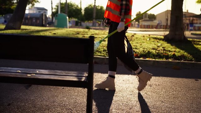 Unrecognizable groundskeeper in reflective safety vest walking through park with rake and garbage bag, preparing for seasonal landscape maintenance and cleanup.